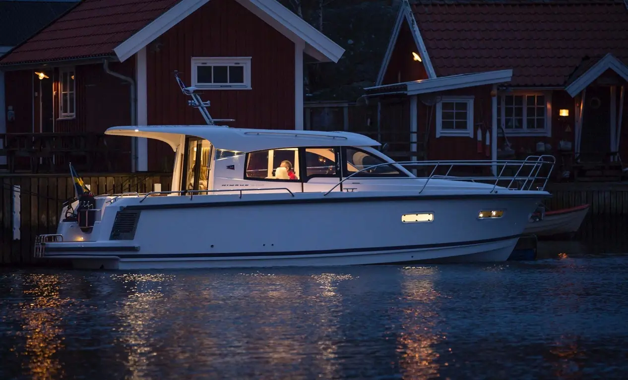 A white motorboat is docked by red wooden houses at dusk, with warm lights reflecting on the water's surface.